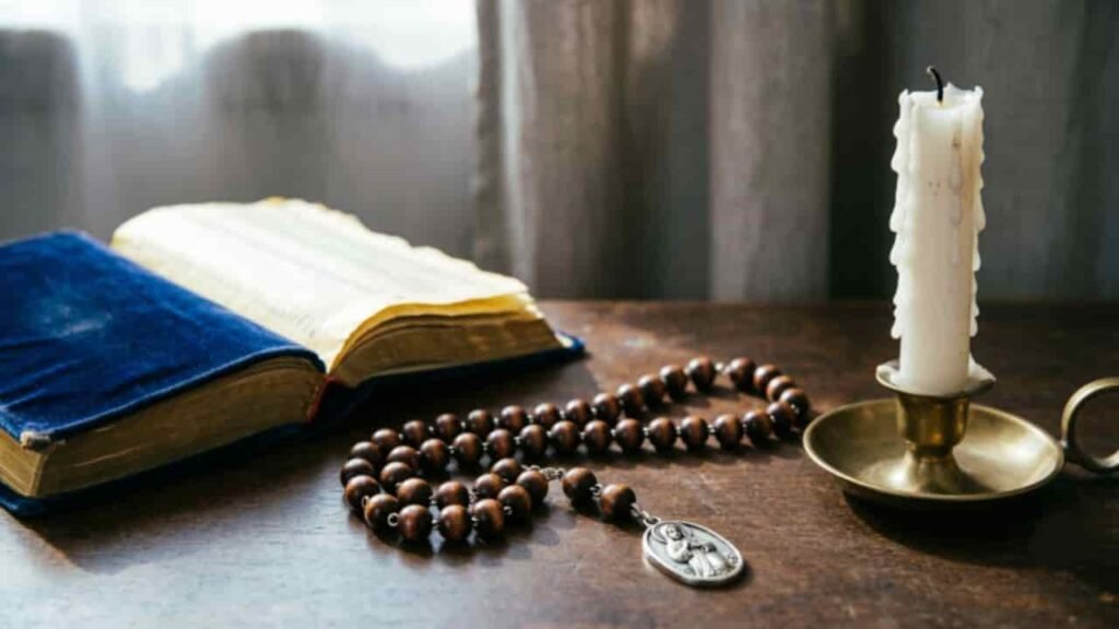 An open prayer book displaying closing Rosary prayers next to wooden rosary beads with a miraculous medal, a burning candle, and white roses on a rustic wooden table.