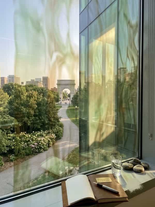 An open leather journal with a fountain pen sits on a window sill alongside glasses of water and smooth river rocks, overlooking a park pathway leading to a stone arch monument.