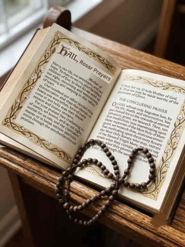 An open prayer book featuring the Hail, Holy Queen prayer alongside a set of wooden rosary beads.