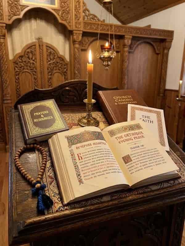 An open Orthodox prayer book featuring evening prayers resting on a carved wooden lectern alongside candles, a prayer rope, and religious resources.